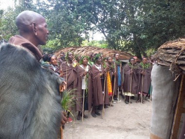 A group of Ogiek people, most of whom are wearing crowns or necklaces made of green leaves, stand in a line in front of a thatched roof building. Their mouths are open as if talking or singing.