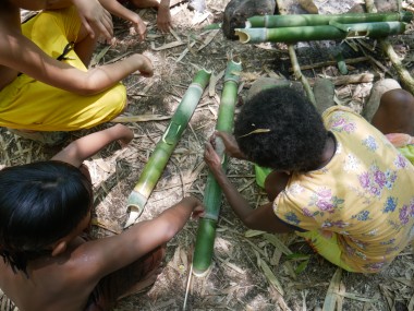 A group of children kneel on the ground around a cluster of green bamboo stalks. Sunlight dapples the children and bamboo.