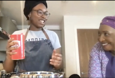 A young Nigerian woman holding a salt shaker above a large bowl, looking to the right at an older Nigerian woman, who is looking back at her and smiling.