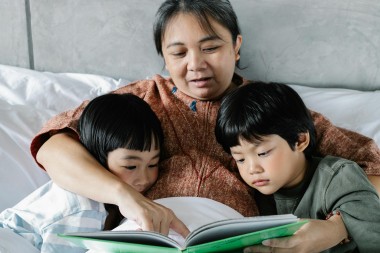A grandmother and two small children cuddled up in bed together. The woman is pointing to a book she is reading, and the children are looking at the book.