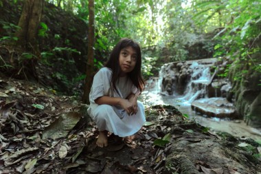Photo of young girl in white dress sitting near a stream in a forest