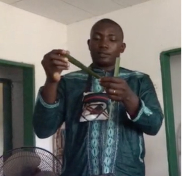 A young man in a green embroidered shirt holds up two partly woven palm fronds
