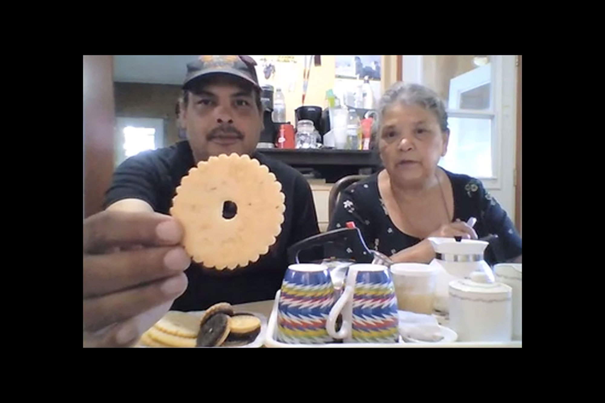 A Zoom screenshot of a man and an older woman, sitting in a kitchen. In front of them on the table are teacups, a tea pot, and a plate of cookies. The man is holding a cookie up to the camera.