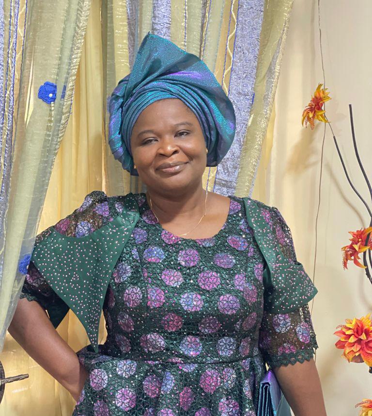 Dr. Bolanle Arokoyo, wearing a teal head wrap and a green and purple dress, smiles in front of a gold-colored curtain.