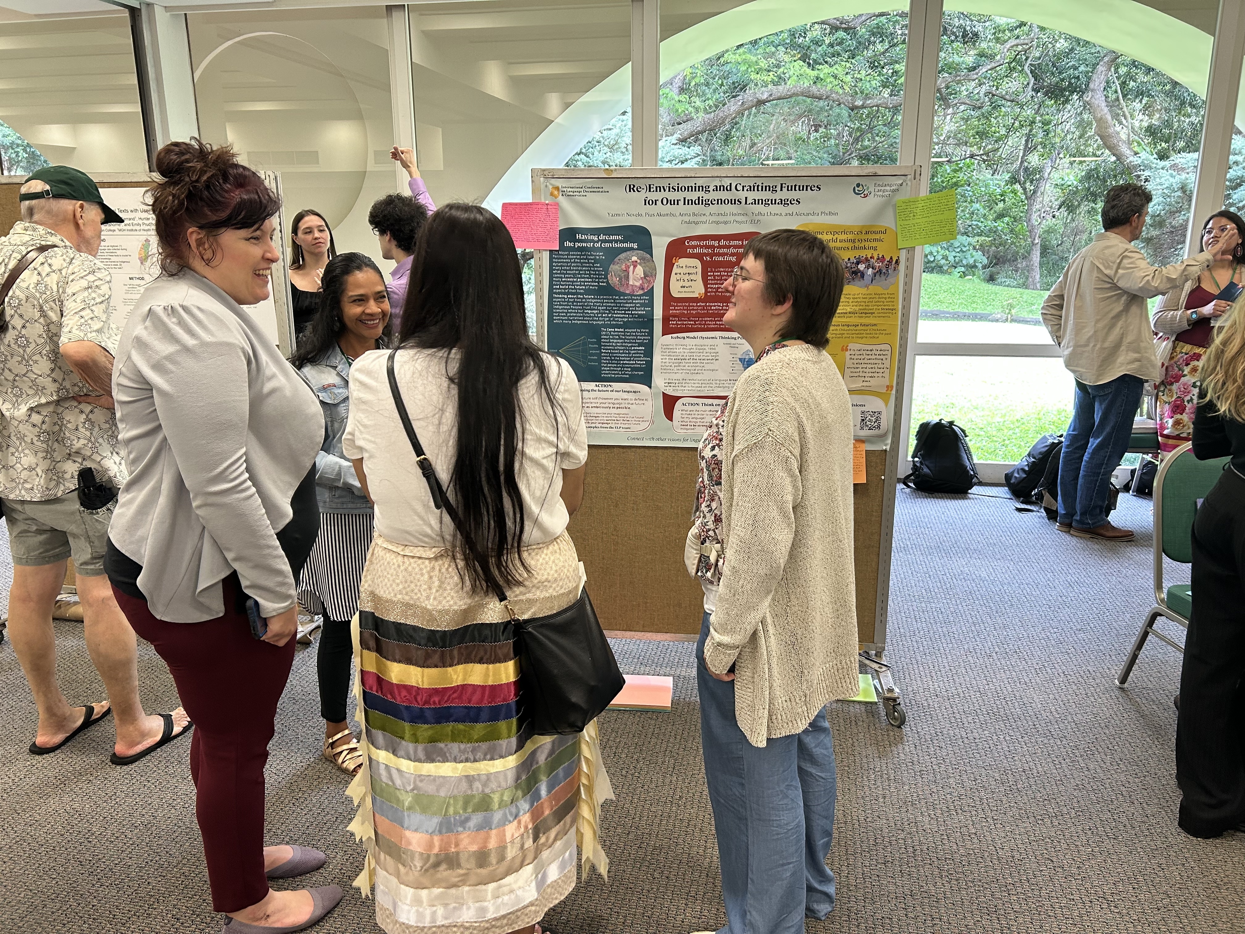 Four women stand in front of a colorful poster in a conference room overlooking a garden.