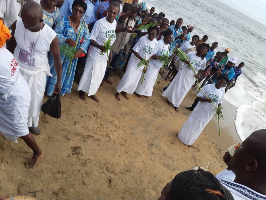 Women wearing white t-shirts and wrap shirts hold palm leaves as they dance in a semi-circle on the beach. Surrounding them is a crowd of people, some wearing patterned blue tunics. A jengú dance, an Iyasa tradition, held at Campo during Joba ja Iyasa (Iyasa Day) celebrations